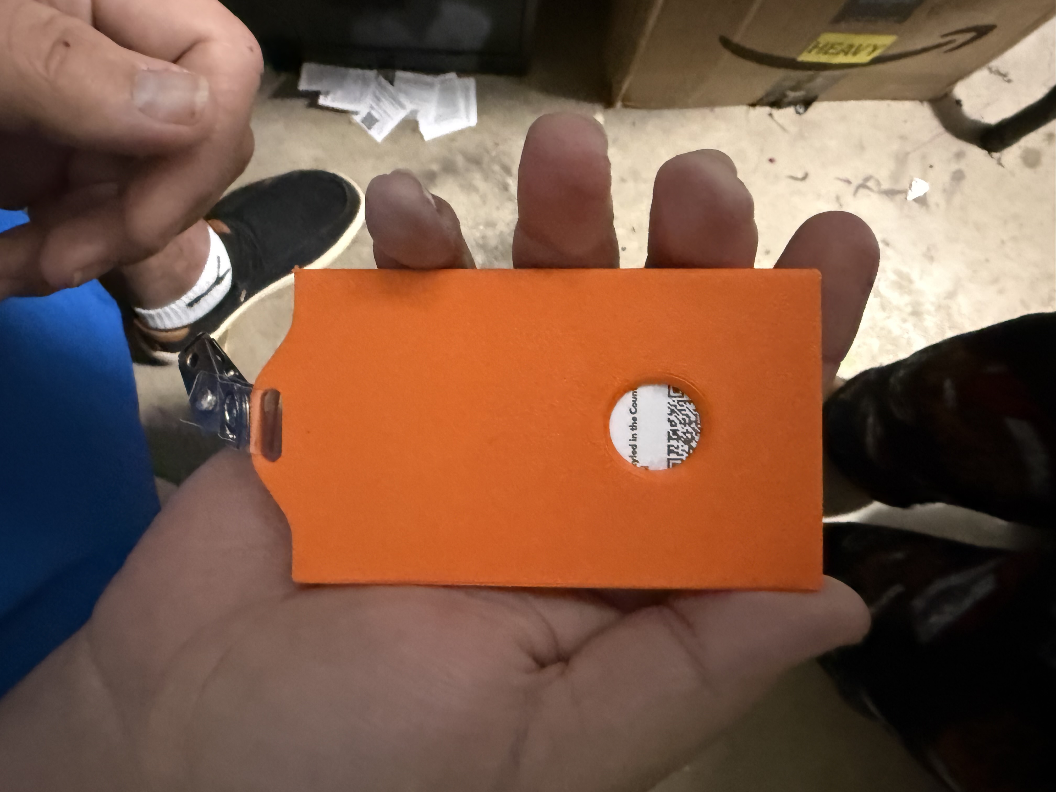 An orange badge holder held in a hand, showing its reverse side, a rectangular attachment slot at the top, and a circular opening near the bottom.