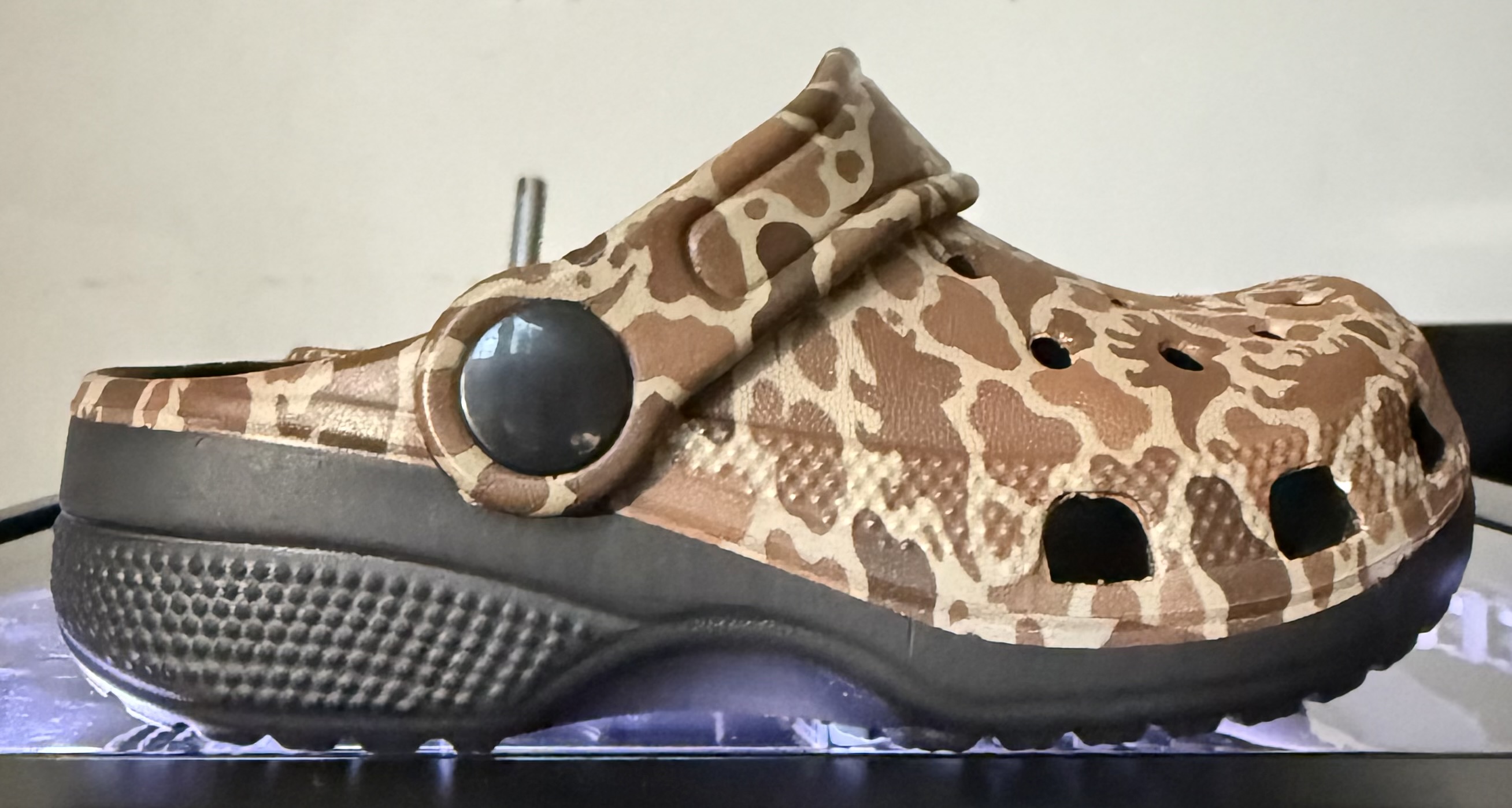 Side profile of one customized Crocs shoe, showing its brown-and-tan dinosaur pattern, strap, black side fastener, and black sole.